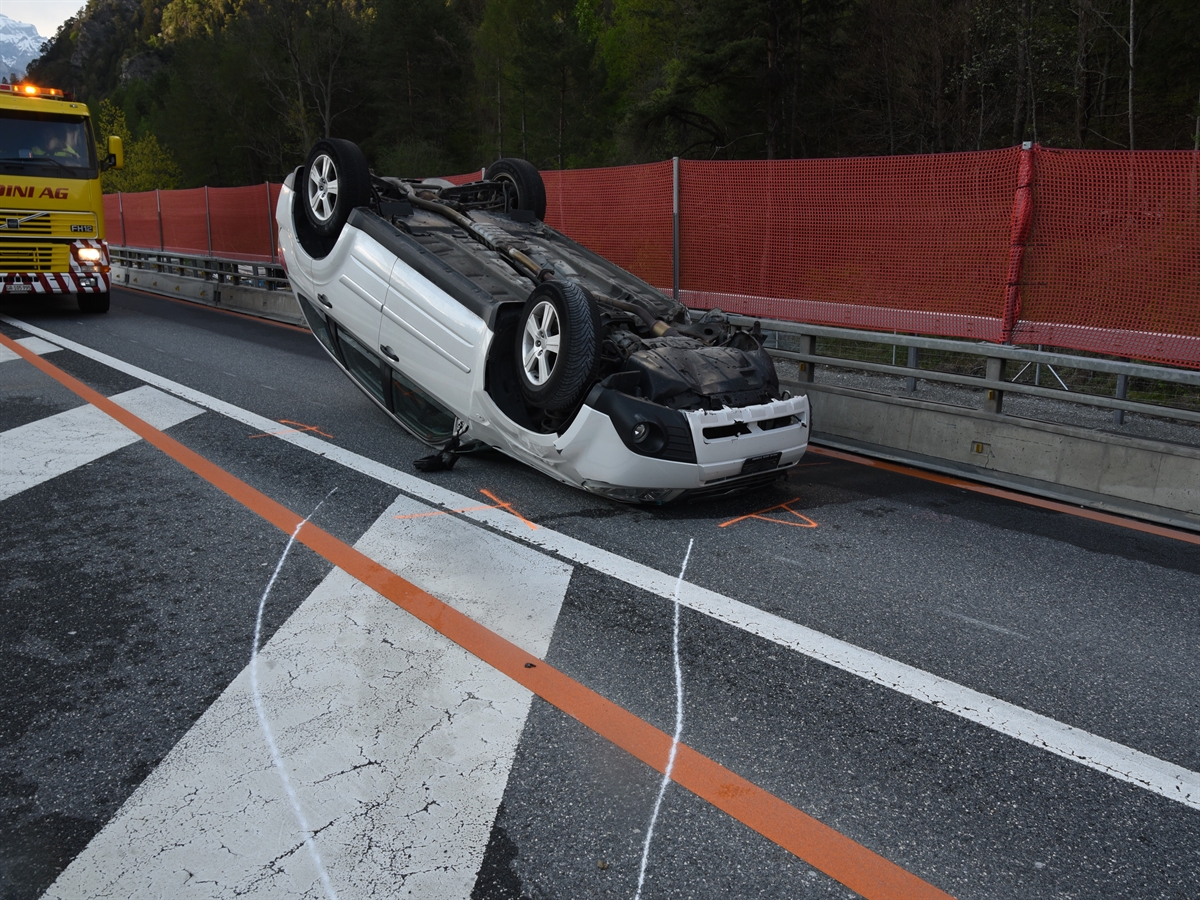 Das Unfallauto, ein weisser Kombi, von schräg seitlich auf dem Dach liegend. Dahinter ein Abschleppfahrzeug.