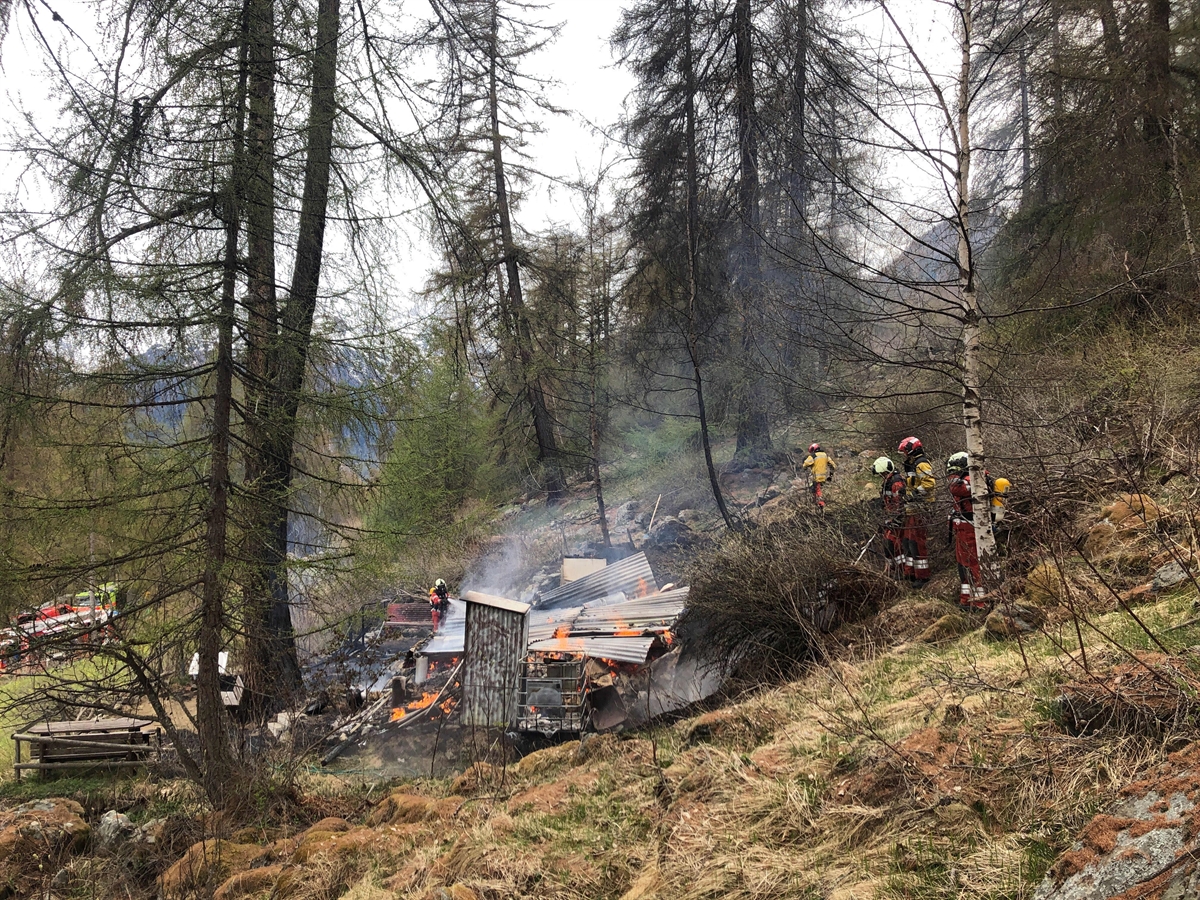 Sicht von der Seite: In der Mitte des Bildes ein Trümmerhaufen der zerstörten Hütte. Einige Feuerwehrleute, die sich um den Brand kümmern. Viele Bäume und Wiese.
