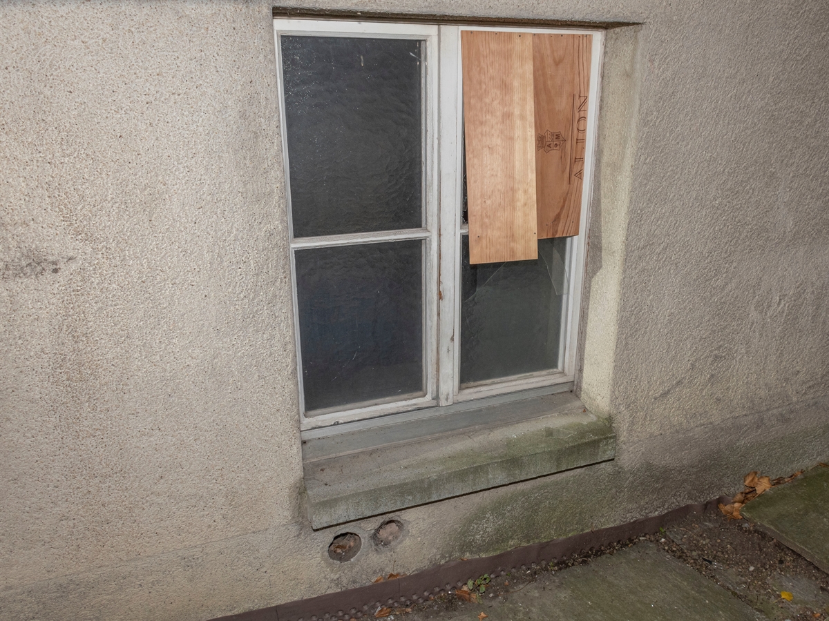 Ein fast bodenebenes Fenster, dessen Glas beschädigt ist. Auf der beschädigten Stelle sind behelfsmässig Holzbretter angebracht.