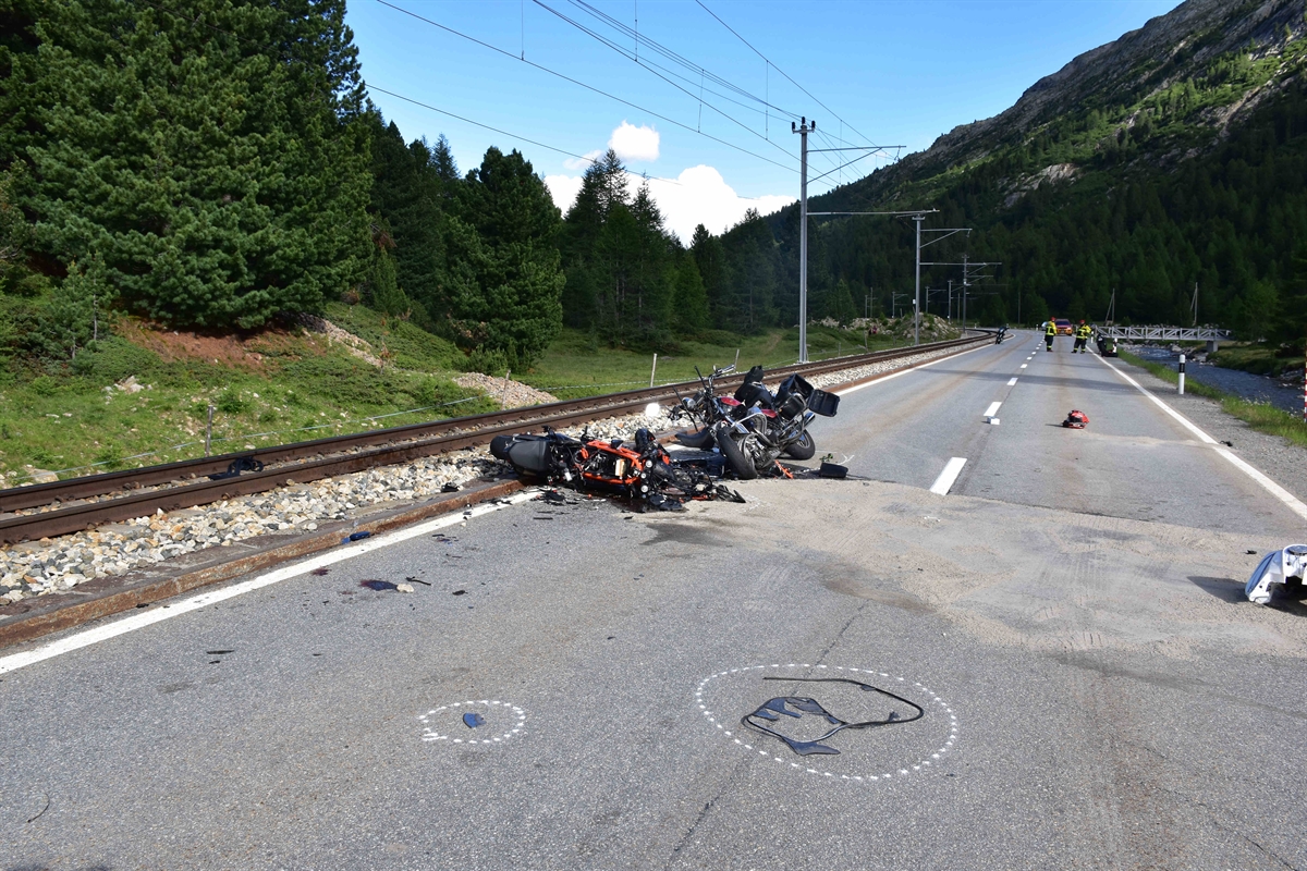 Übersichtsbild der Unfallstelle. Sicht auf die Strasse in Richtung Bernina. Linksseitig sind die Geleise, die beiden total beschädigten Motorräder liegen gleich daneben. Auf der Strasse sind verschiedene Teile verstreut. Unter anderem ein Helm und Teile der Motorräder.