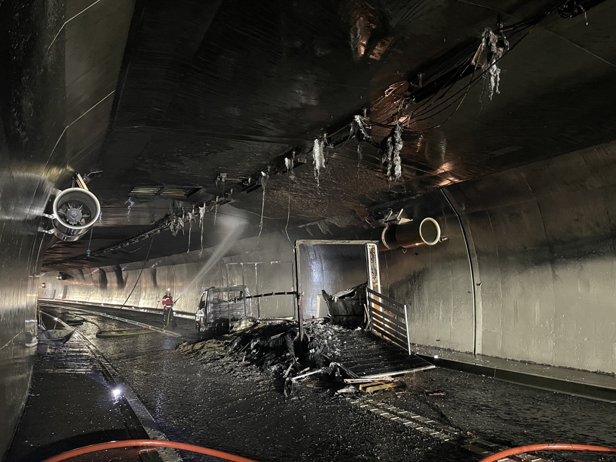 Aufnahme der abgebrannten Fahrzeugkombination. Von der Tunneldecke hangen beschädigte technische Installationen runter. Ein Feuerwehrmann, welcher mit Spritzwasser die Tunneldecke bewässert.