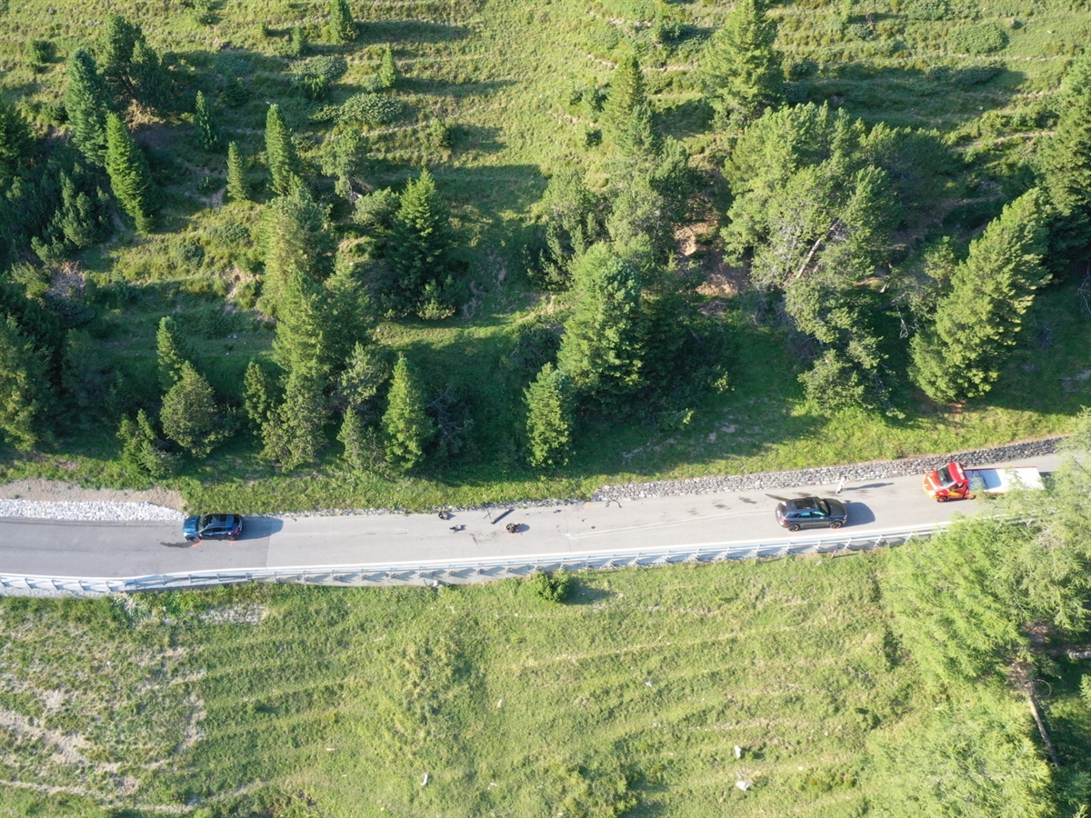 Die Drohnenaufnahme zeigt links auf der quer durchs Bild verlaufenden Strasse das talwärts und rechts das bergwärts gefahrene Auto. Oben an der Strasse ansteigendes und unten an der Strasse abfallendes Wiesland.