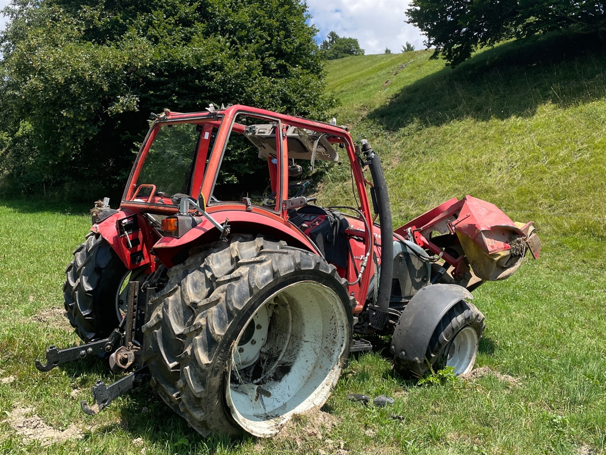 Der total beschädigte Traktor am Fusse eines Abhangs. Zwischen Laubbäumen hindurch führt eine Spur mit beschädigtem Terrain zum Fahrzeug.