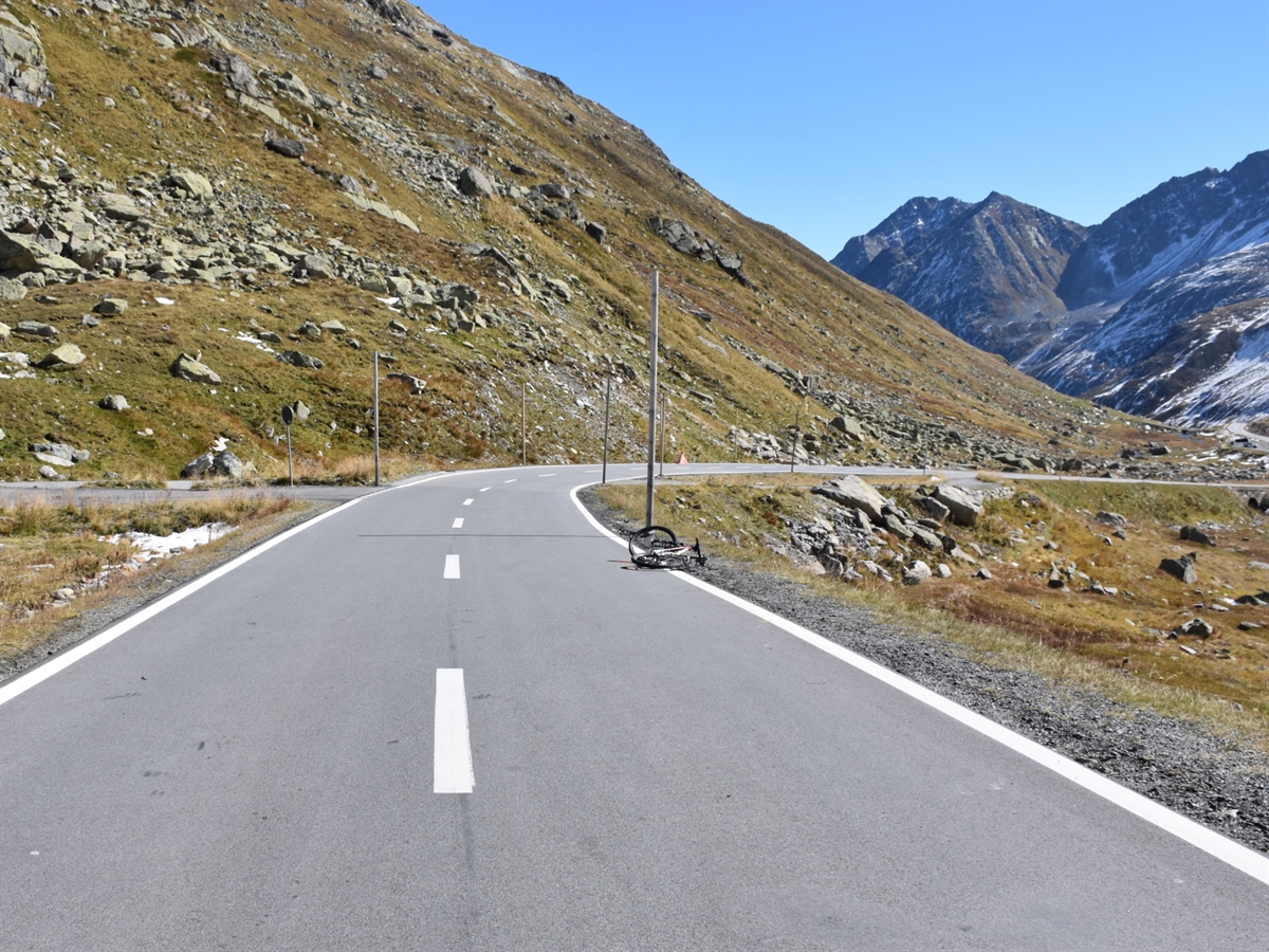 Die kurze Gerade, welche in eine Rechtskurve übergeht. Am linken Strassenrand liegt das Fahrrad. Im Hintergrund Berge, die den blauen Horizont abgrenzen.