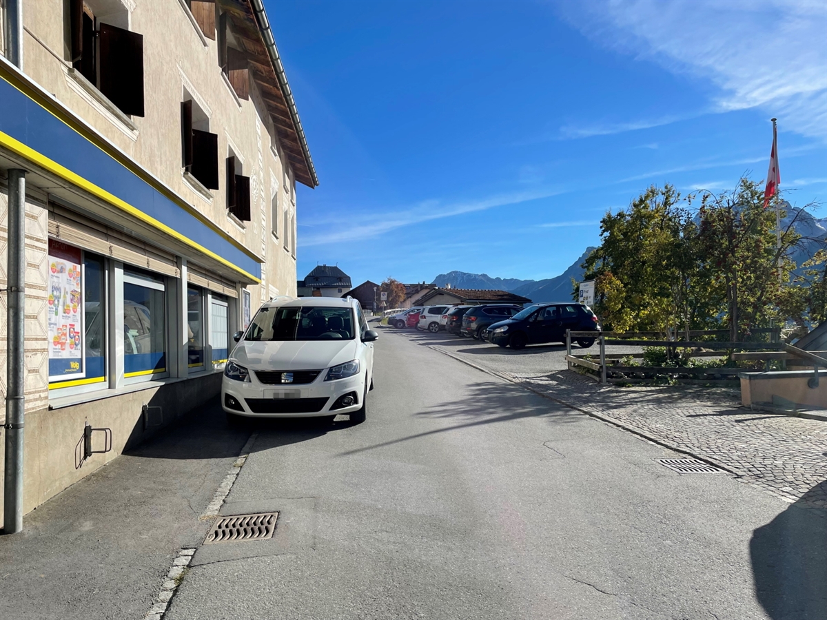 Die Dorfstrasse von Ftan inmitten der Engadiner Bergwelt. Rechtsseitig der Strasse der Parkplatz und linksseitig ein Lebensmittelgeschäft. Der blaue Engadiner Himmel rundet das Bild ab.