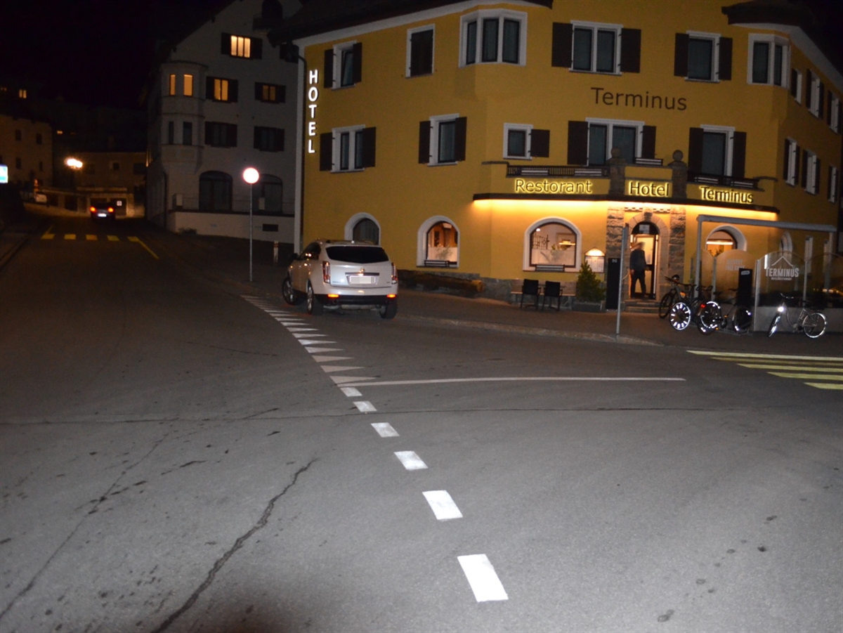 Aufnahme in der Dunkelheit. Eine Kreuzung, im Zentrum ein gelbes Gebäude und ein auf der Strasse abgestelltes weisses Auto.