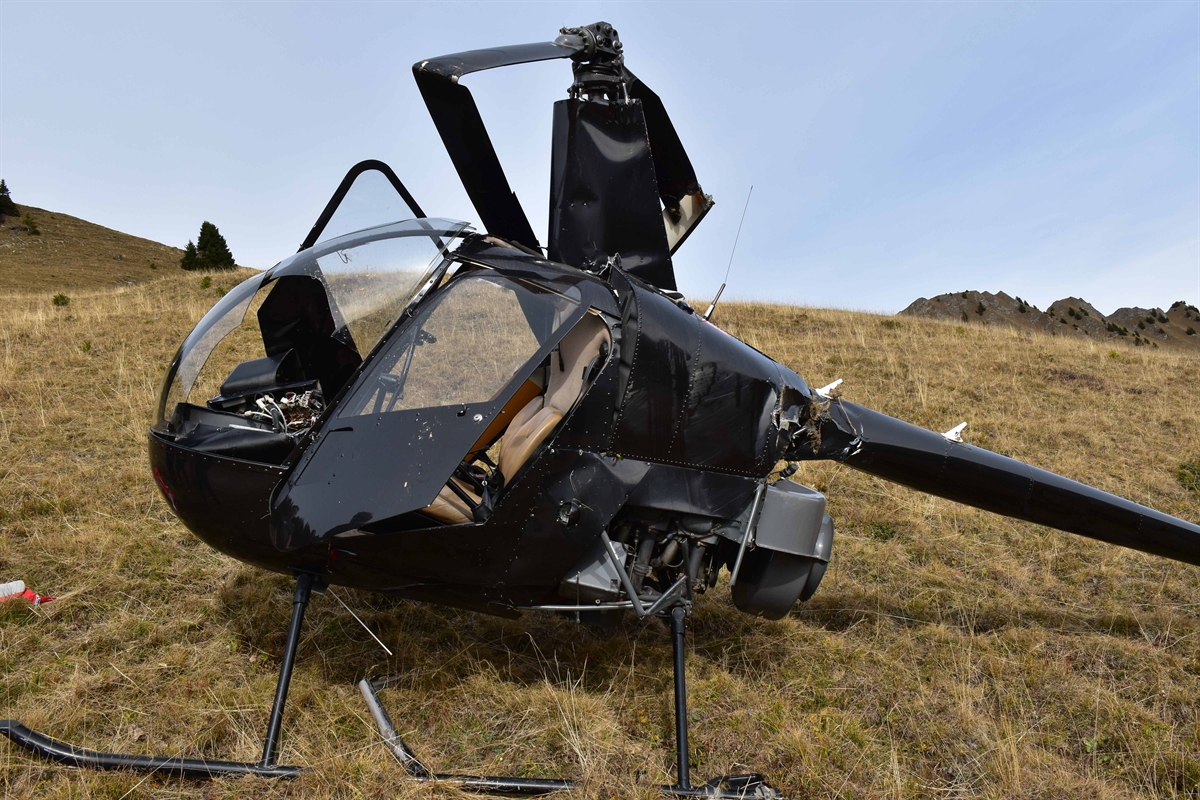 Schwarzer Helikopter auf steilem Wiesland. Die Frontscheibe ist zerschlagen, die Türen sind offen. Die Rotoren sind komplett gebrochen und hängen runter. Verschiedene Beulen und Beschädigungen sind zu sehen.