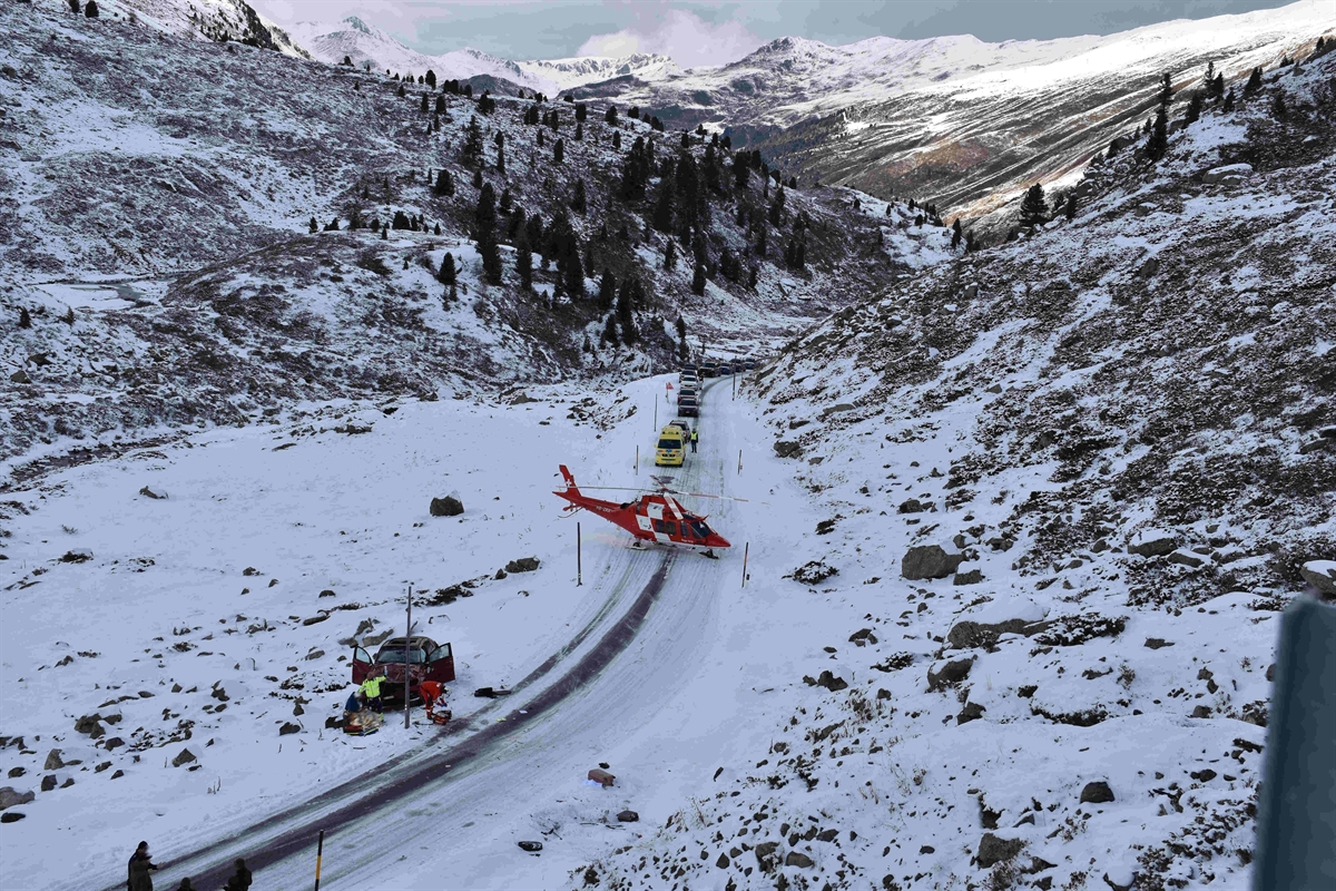 Das Unfallauto steht unterhalb der schneebedeckte Flüelapassstrasse. Der Regaheli steht mitten auf der Strasse und die Ambulanz dahinter. Hinter der Ambulanz steht eine Fahrzeugkolonne.