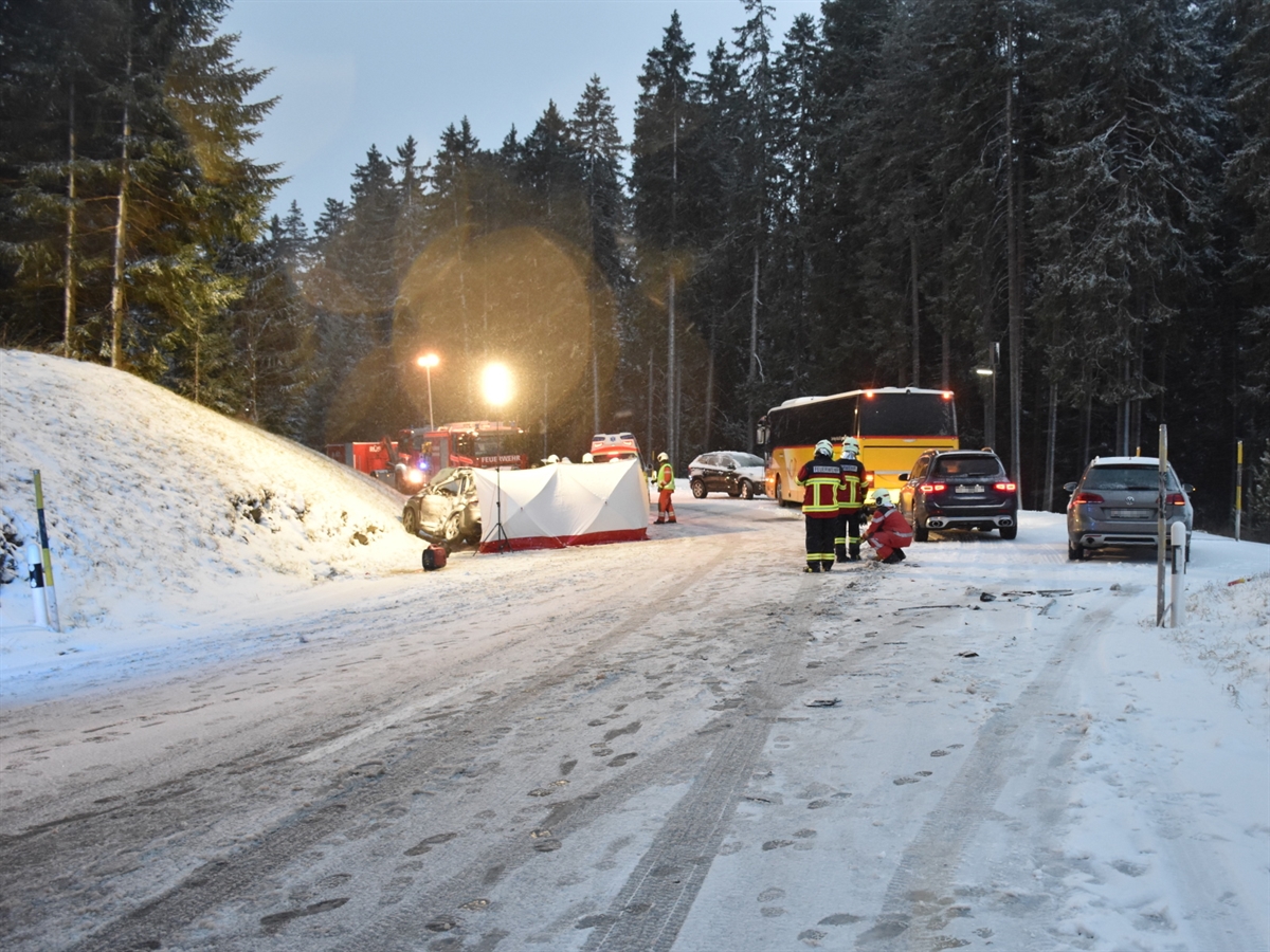 Rechts auf der schneebedeckten Strasse zwei Autos und entfernter das Postauto von hinten. Links das von einer Plane vor Sicht geschützte Unfallauto und Einsatzkräfte sowie ein Feuerwehr- und ein Ambulanzfahrzeug