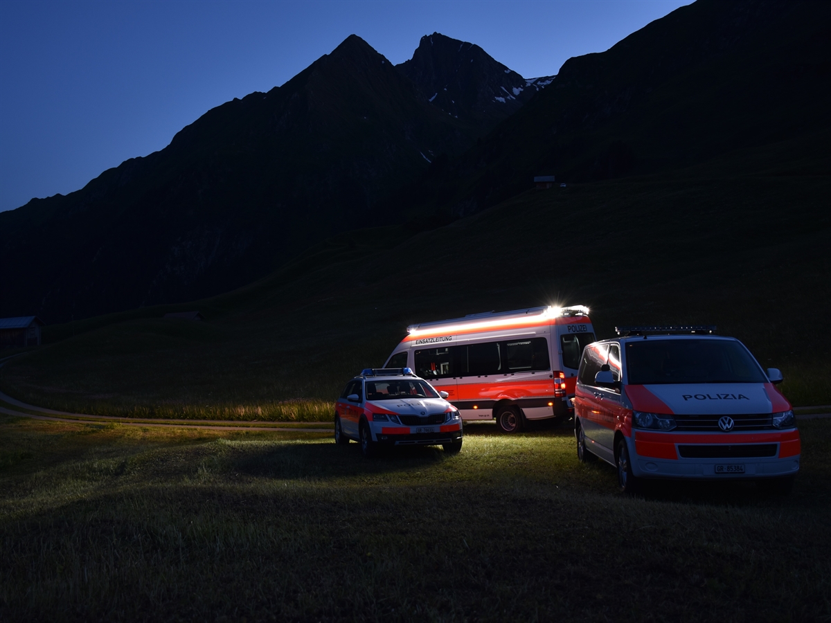 Aufnahme in der Dämmerung. Drei Einsatzfahrzeuge der Polizei stehen auf einer Wiese. Eines beleuchtet die nahe Umgebung. Im Hintergrund heben sich Berge vom wolkenlosen Himmel ab.