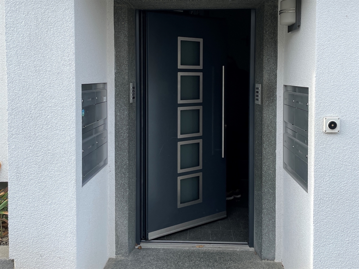 Eine Treppenhaustüre von aussen. Sie steht halb offen. Links und rechts an den Wänden sind Briefkästen. 