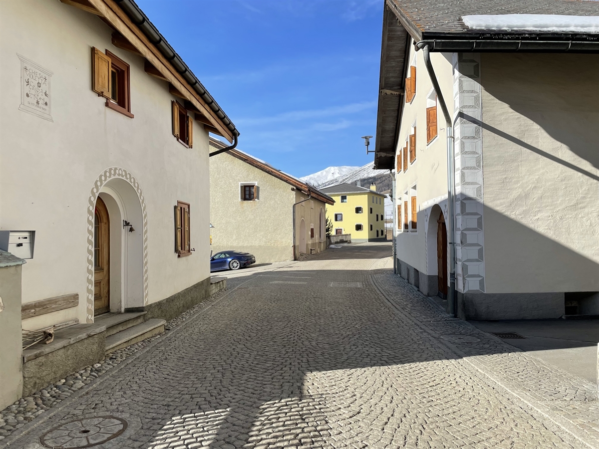 Die gepflasterte Dorfstrasse von S-chanf wird mit typischen Engadinerhäusern begrenzt. Der blaue Himmel rundet das idyllische Bild ab.