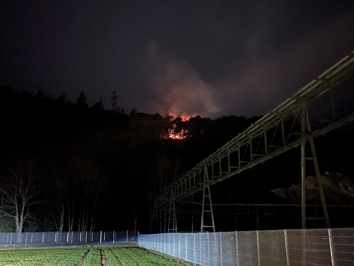 Nachtaufnahme aus einem umzäunten Gelände auf den Brandort hoch. 