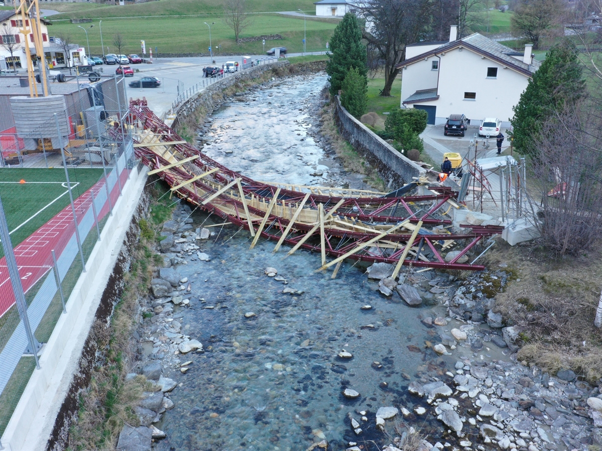 Luftaufnahme auf die im Fluss Poschiavino liegende eingestürzte Brücke. Links des Flusses ein Fussballplatz, rechts des Flusses ein Haus.