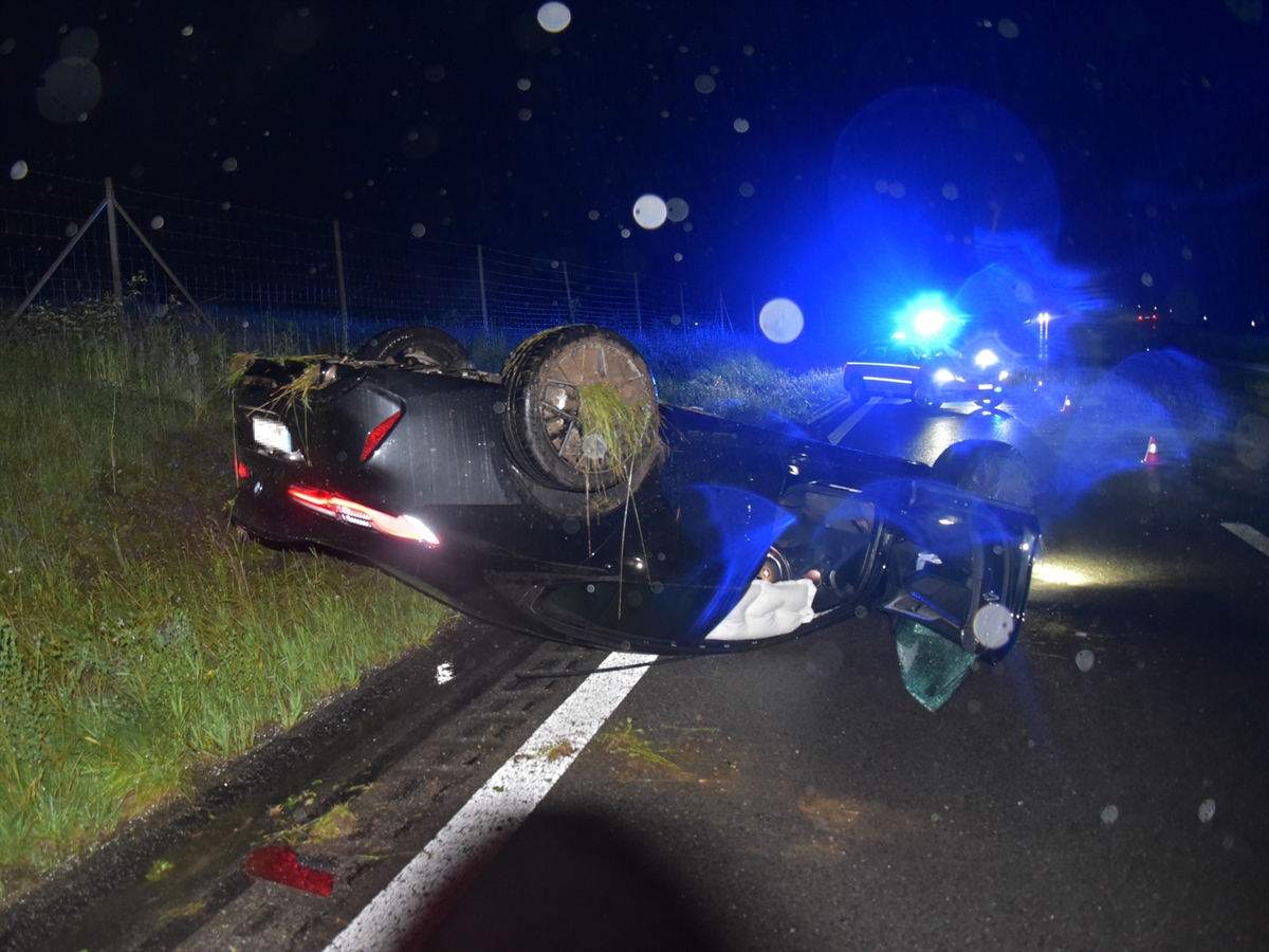 Das Unfallauto liegt auf dem Dach auf der Fahrbahn der Autobahn A13 neben dem Erdwall. Hinter dem Unfallauto erhellt das Blaulicht von einem Patrouillenwagen der Polizei die regnerische Nacht.