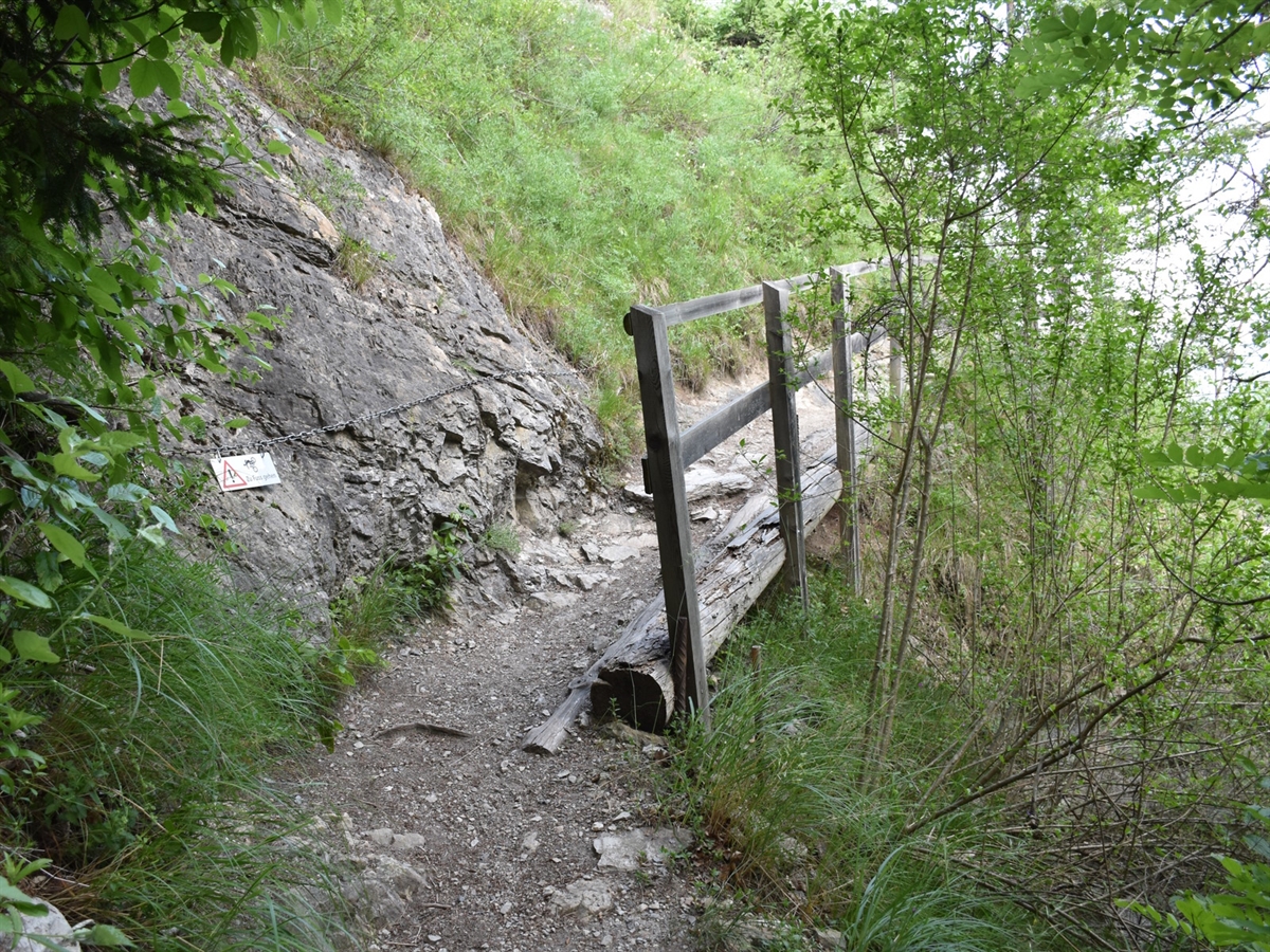 Ein schmaler und unbefestigter Weg, rechts abschnittweise durch einen Metallzaun vom dortig abfallenden Gelände abgegrenzt wird.