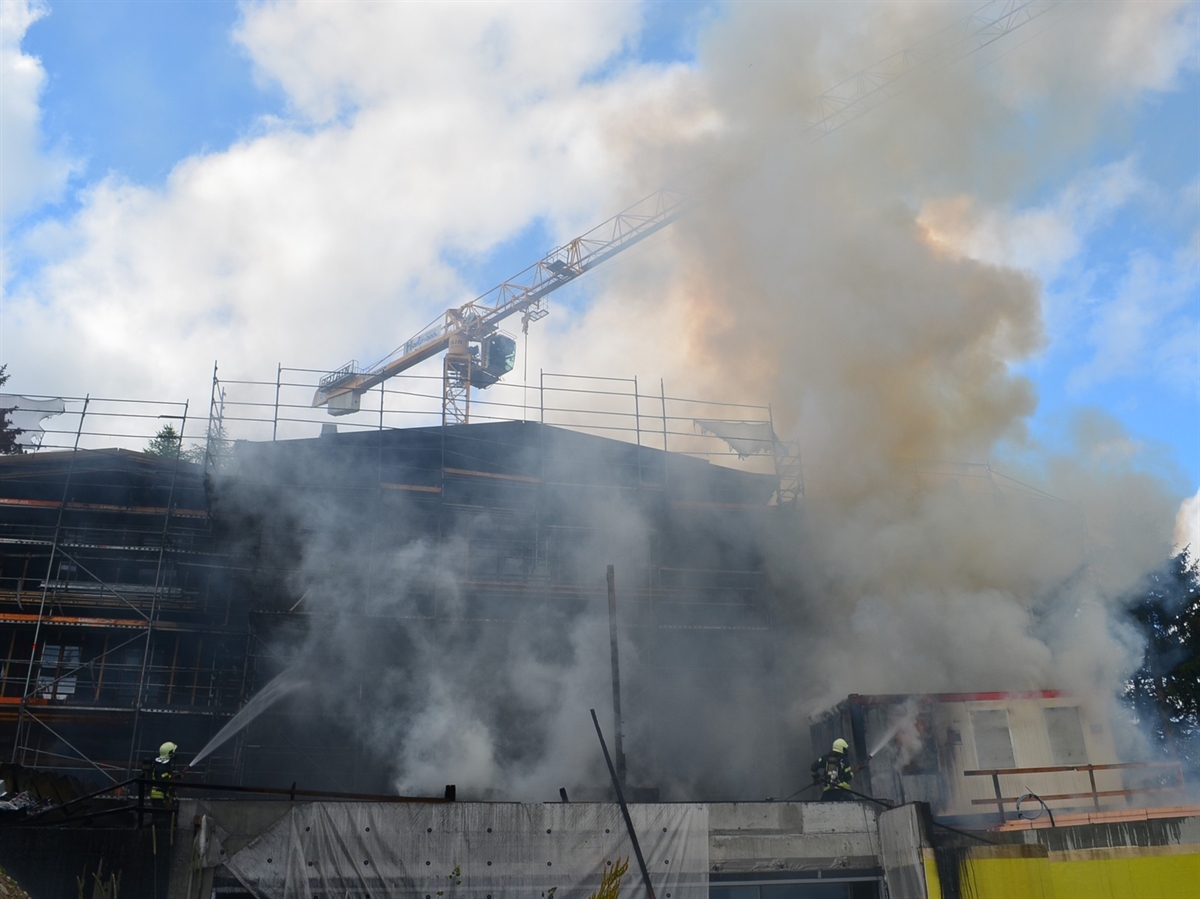 Im Vordergrund Feuerwehrleute bei den Löscharbeiten am Objekt. Vor diesem steigt Rauch auf, im Hintergrund leicht bewölkter Himmel.