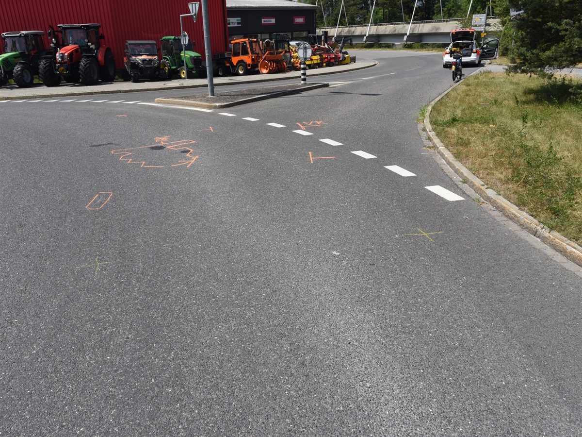 Im Kreisel orange markierte Unfallspuren. Rechts im Kreiselausgang ein Mofa und ein Polizeiauto. Links entlang einer roten Fassade aufgereihte Landmaschinen.