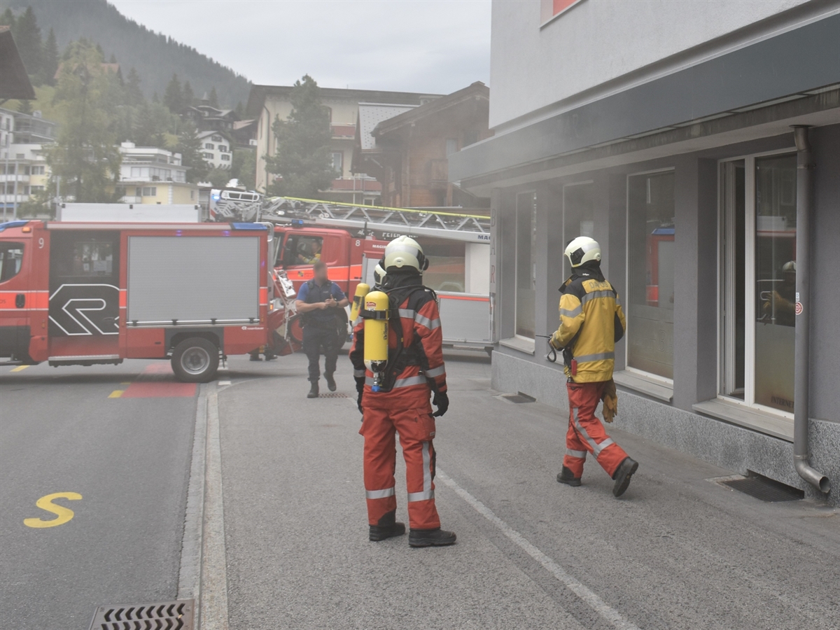 Vor einem Gebäude im starken Rauch zwei Feuerwehrleute, ein Polizist und zwei Einsatzfahrzeuge der Feuerwehr Davos