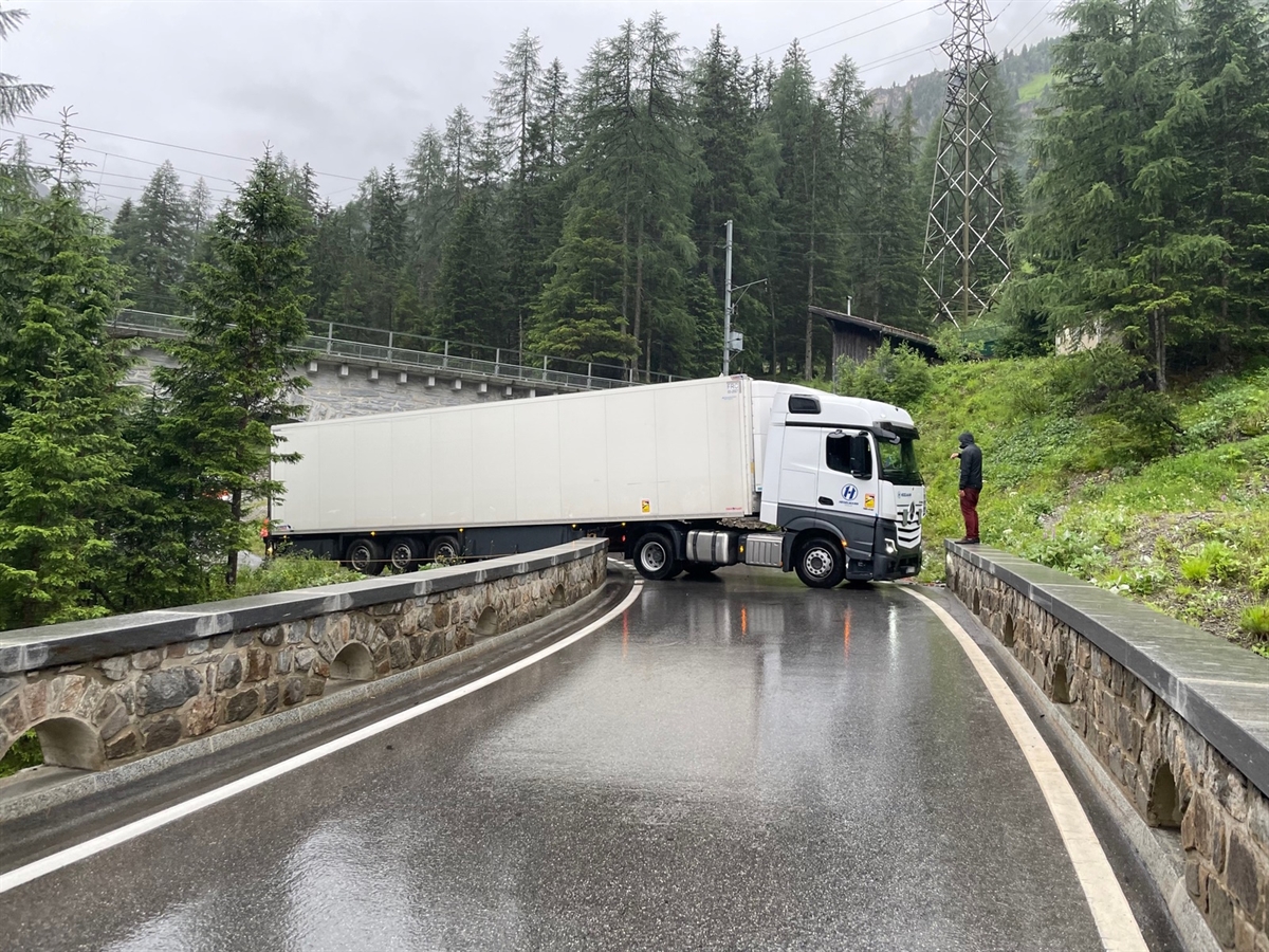 Die Strasse ist durch halbhohe Steinmauern links und rechts begrenzt. Quer auf dieser steht der vordere Teil des weissen Sattelmotofahrzeugs. Rechts eine Hilfsperson, im Hintergrund ein Teil des Bahnviadukts.