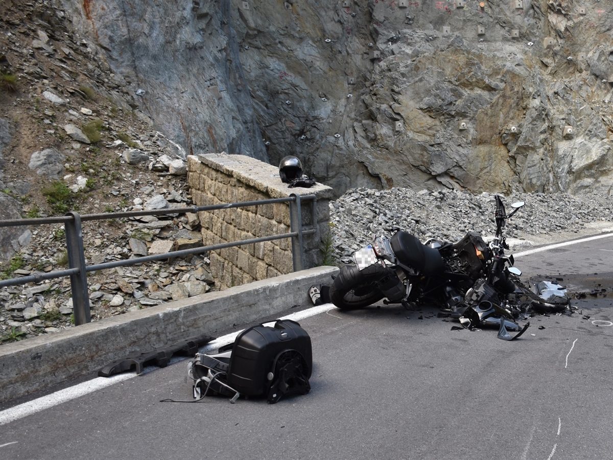 Von schräg hinten das stark beschädigte Motorrad auf der rechten Seite liegend am linken Strassenrand. Im Hintergrund eine Felswand.