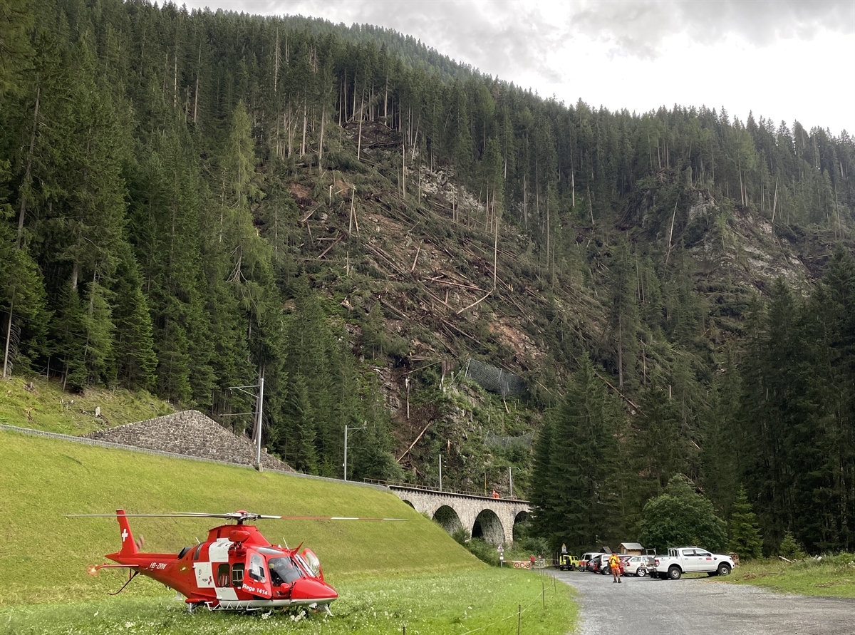 Rega auf Wiese gelandet, im Hintergrund umgeknickte Bäume im Hang