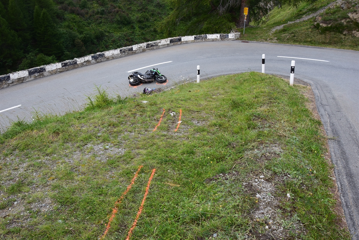 Das Foto zeigt eine Übersichtsaufnahme der Unfallstelle. Man sieht den Strassenverlauf, eine Spitzkehre nach links, in der Mitte das Wiesland und darin sind die Spuren mit rot eingezeichnet, wo das Motorrad abgestürzt ist. Das Motorrad liegt auf der Strasse nach der Kurve.