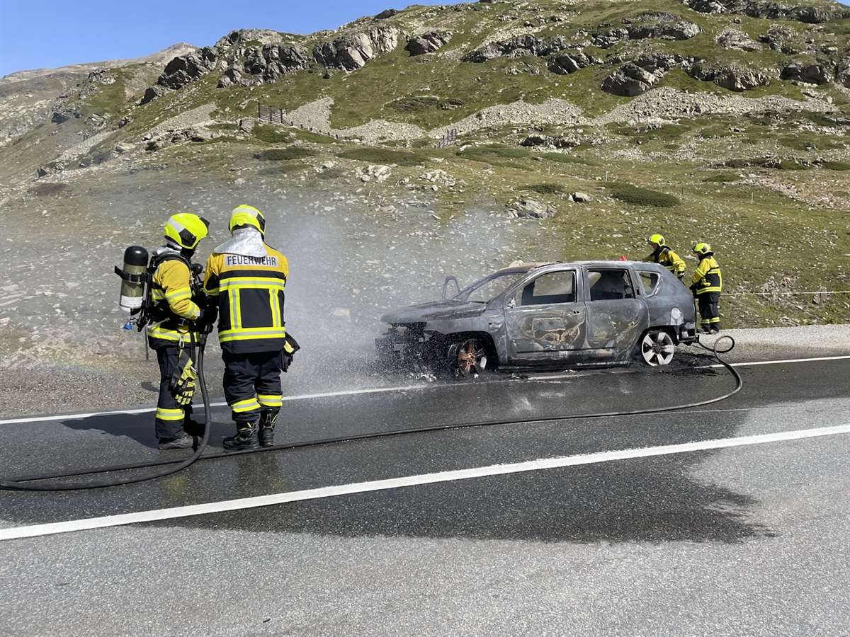 Vier Feuerwehrleute kühlen das vollständig ausgebrannte Auto mit Wasser ab. Umrahmt wird das Bild von einer herrlichen Bergwelt mit stahlblauem Himmel.