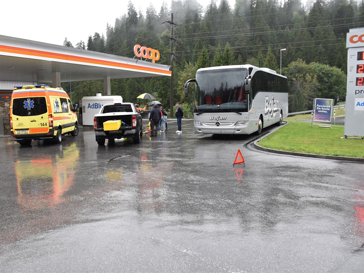 Tankstellenareal. Rechts ein Reisebus, links davon Personen, das Unfallauto und ein Rettungswagen.
