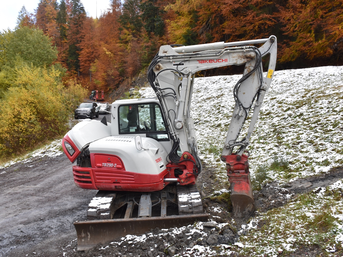 Der beschädigte Bagger steht auf einer leicht verschneiten Wiese. Im Hintergrund stehen zwei Autos sowie ein Traktor mit Anhänger in der farbenprächtigen Landschaft von Versam.