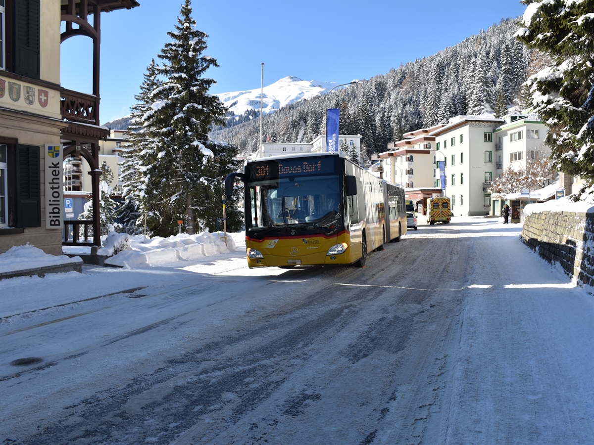 Die schneebedeckte Strasse mit dem Linienbus (Postauto) von schräg vorne. Im Hintergrund Winterlandschaft und blauer Himmel. 