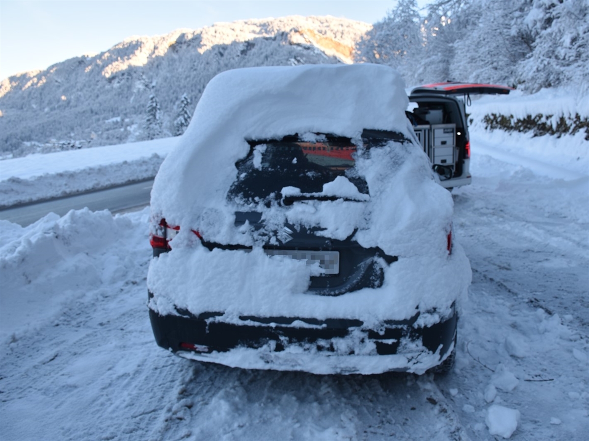 Das Auto von hinten. Die Rückleuchten sind kaum erkennbar und auf dem Dach liegt ca. 30 cm Schnee. Rechts ein Polizeiauto sowie  im Hintergrund tief eingeschneite Landschaft und blauer Himmel.