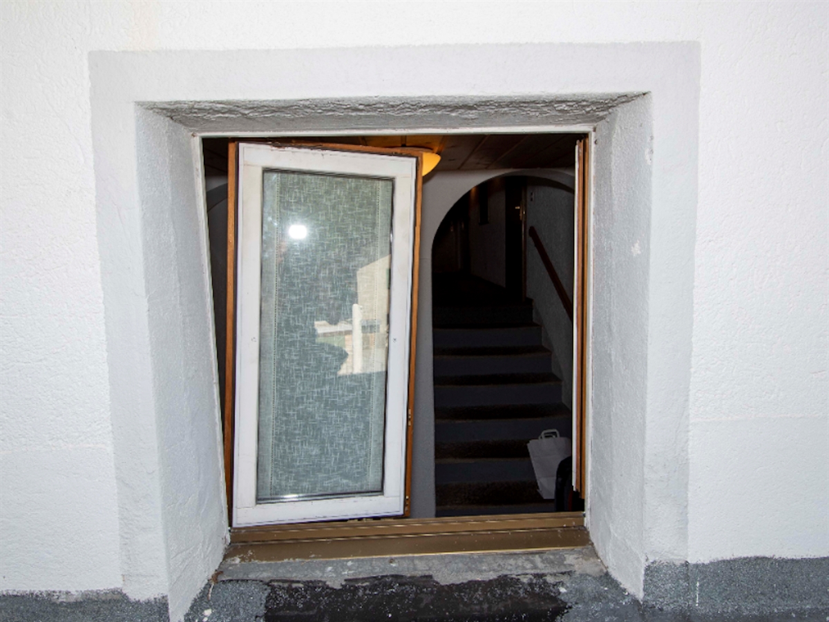 Blick von ausserhalb auf ein mit Gewalt aufgebrochenes Doppelfenster. Im Innern eine nach oben führende Treppe.