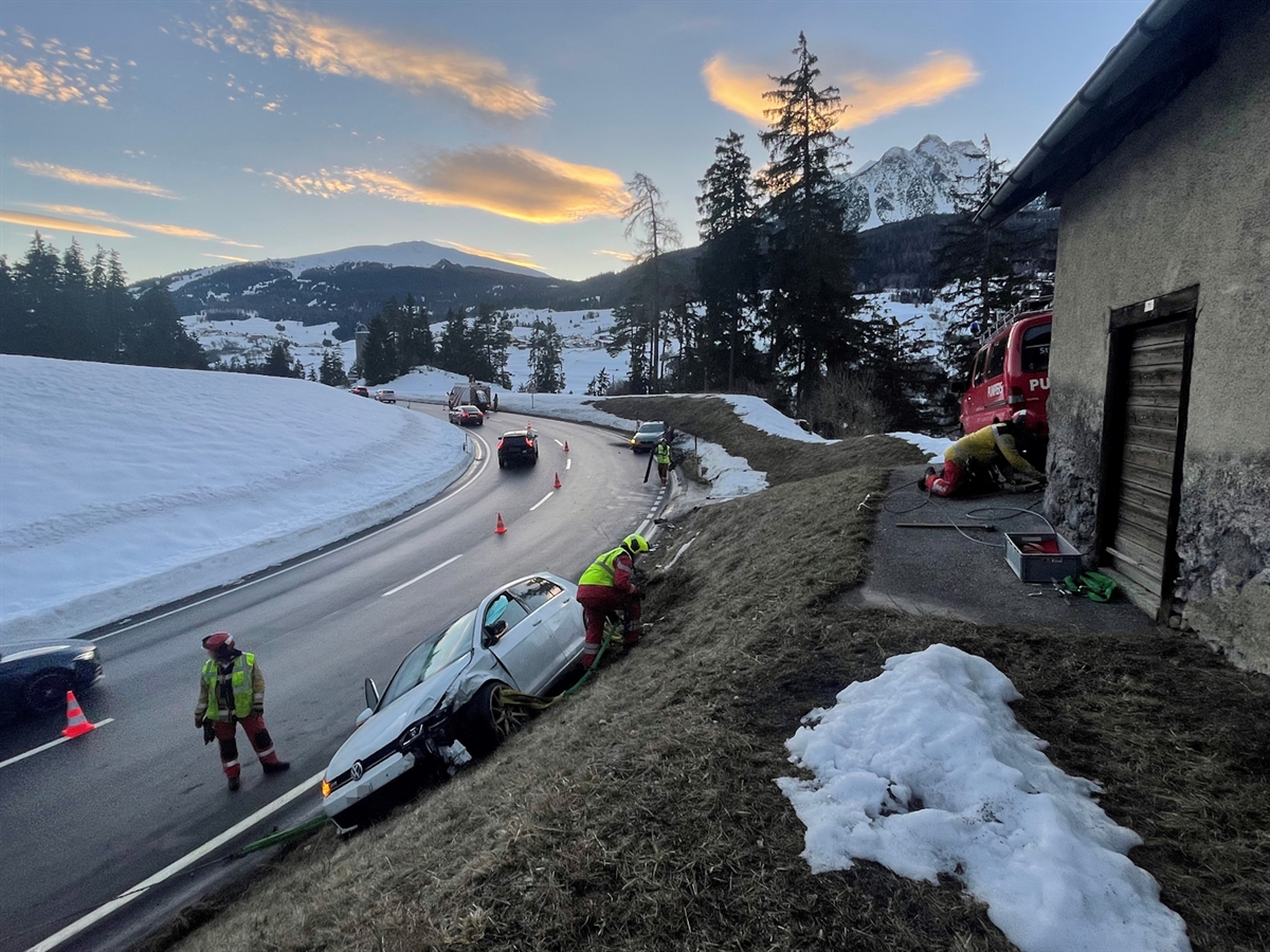 Die Unfallörtlichkeit erstrahlt in abendlicher Dämmerung. In der Bildmitte sichern Feuerwehrleute das Auto in der Böschung. Im Hintergrund das zweite Unfallfahrzeug sowie Fahrzeuge der Polizei und Feuerwehr