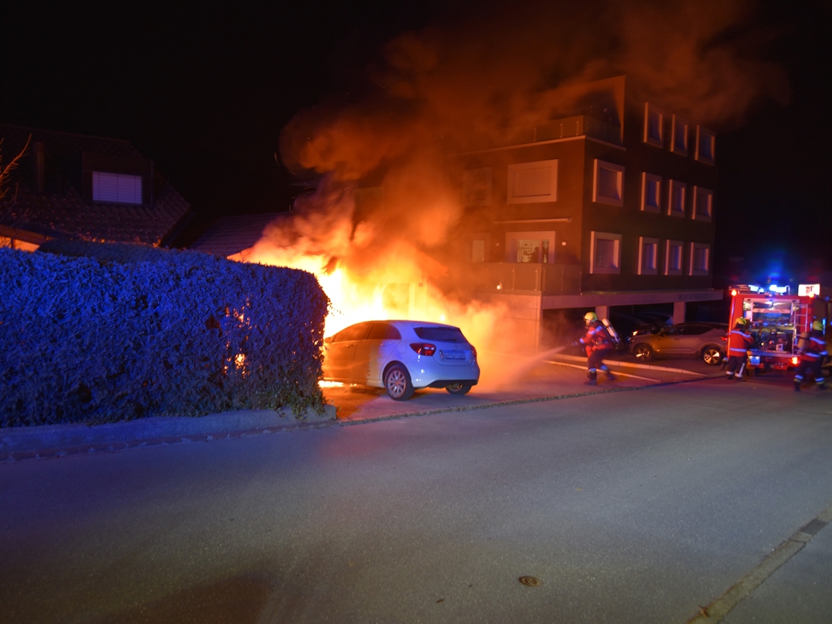 Das Auto von schräg links hinten. Es steht mit brennender Front nahe einer Fassade. Rechts davon Einsatzkräfte der Feuerwehr.