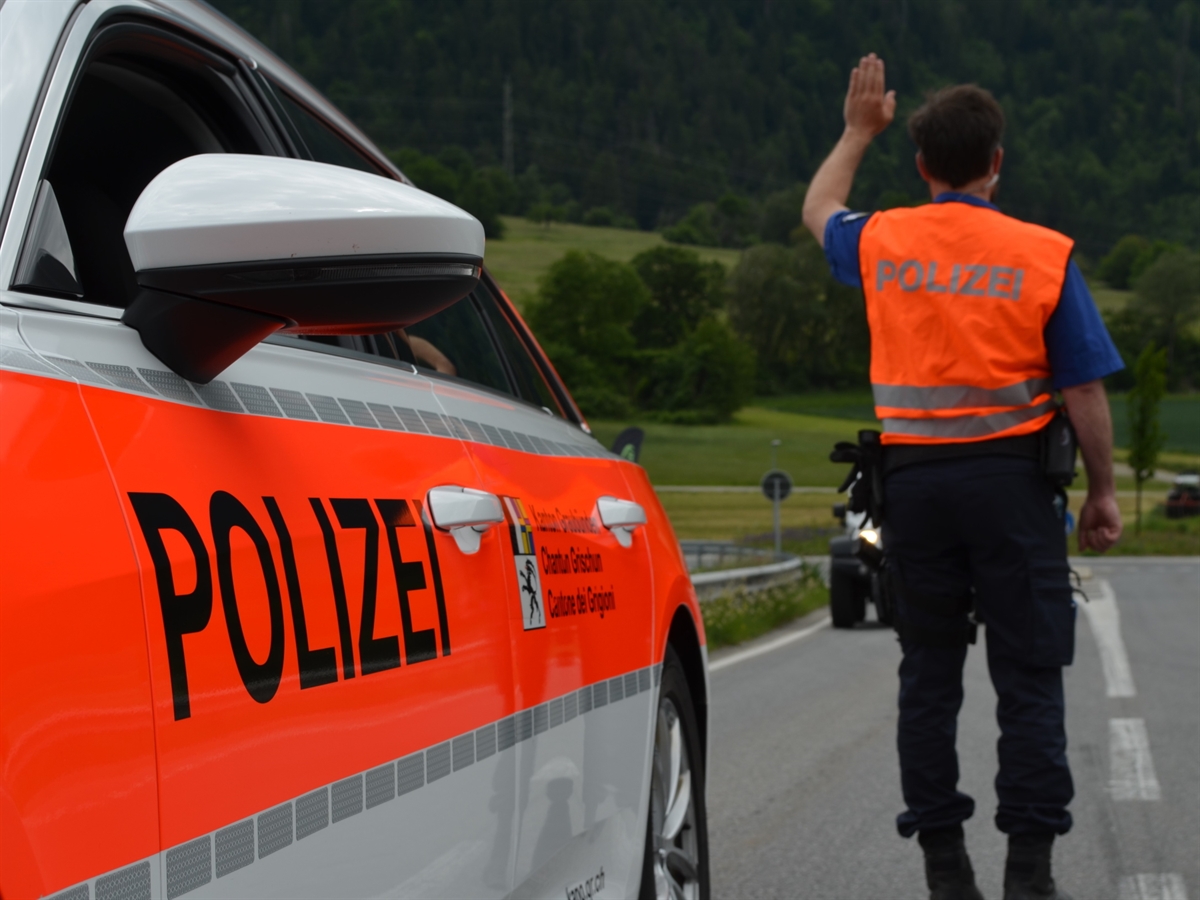 Ein Polizist neben einem Patrouillenfahrzeug der Kantonspolizei Graubünden welcher ein Haltezeichen gibt.