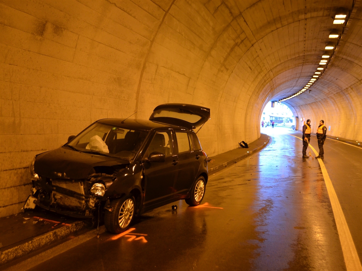 Links an der Tunnelwand das frontal stark beschädigte Auto. Im Hintergrund zwei Polizisten und der Blick aus dem Tunnel ins Freie.