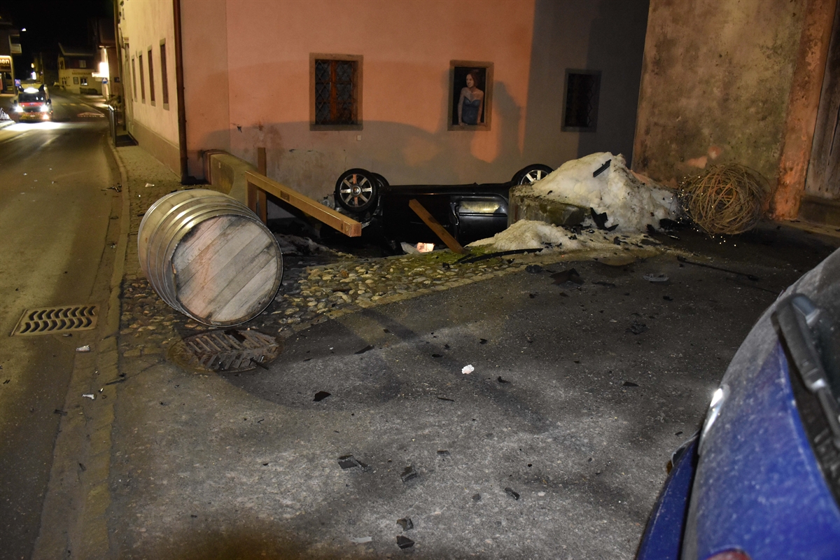 Das Unfallfahrzeug liegt auf dem Dach in einer Seitengasse. Die Räder in der Luft sind ersichtlich. Rechts ist das Heckteil des parkierten Fahrzeuges. An der Hausfassade ist der Schaden ersichtlich. Überall liegen Teile herum.
