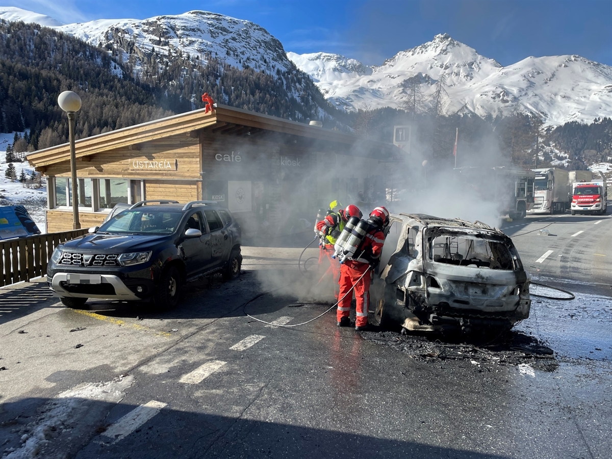 Das ausgebrannte Auto, das parkierte und angesengte Auto und daneben Einsatzkräfte. Dahinter ein Feuerwehrauto sowie Winterlandschaft mit blauem Himmel.