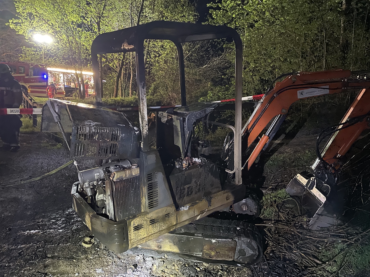 Die völlig ausgebrannte Kabine des Kleinbaggers. Im Hintergrund ein Feuerwehrfahrzeug sowie Feuerwehrleute im hell ausgeleuchteten Wald