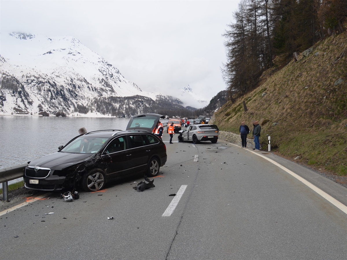 Im Vordergrund ein frontal und seitlich stark beschädigtes Auto von vorne, links der Silsersee. Im Hintergrund ein ebenfalls vorn links stark beschädigtes Auto von hinten.
