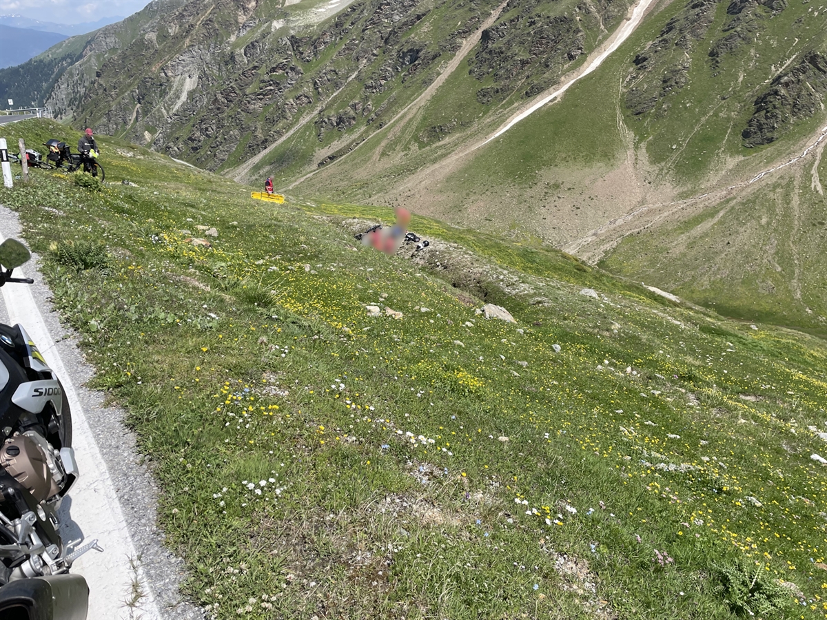 Am Bildrand die Umbrailstrasse mit Blick über den Abhang hinunter. In einem Graben leisten Einsatzkräfte mit Drittpersonen Erste Hilfe beim Verunfallten.