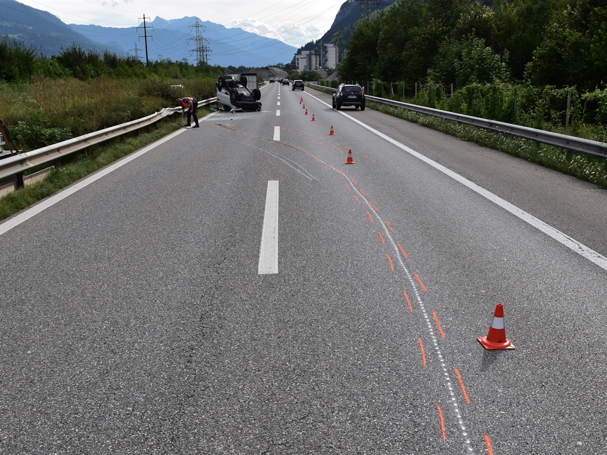 Im Vordergrund Schleuderspuren, die nach links zur Mittelleitplanke verlaufen. Dort liegt das Zugfahrzeug auf seinem Dach. Im Hintergrund Berge.