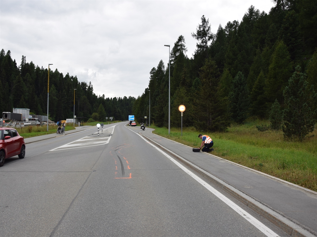Bremsspur auf der Engadinerstrasse H27. Auf dem Trottoir kniet ein Polizist bei einem Koffer. Im Hintergrund sieht man ein Motorrad sowie ein Polizeifahrzeug auf einem Ausstellplatz stehen