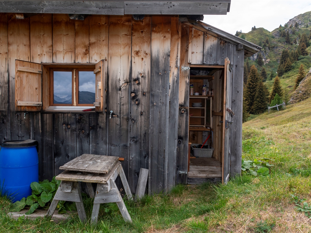 Ein Maiensäss mit einem seitlichen Vorbau bei welchem die Holztüre aufgebrochen ist. Im Hintergrund Berglandschaft.