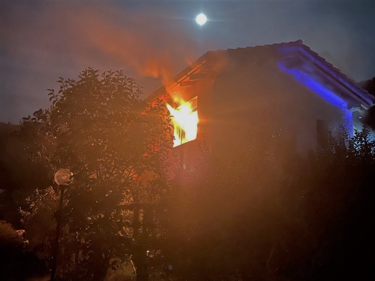 Ein Haus von schräg unten. Aus einem Fenster im obersten Stock dringen Flammen nach aussen.