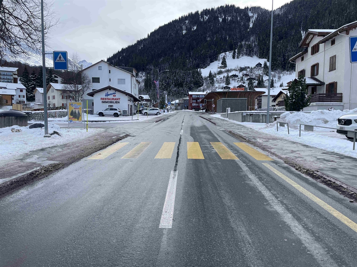 Standpunkt von der Strassenmitte ein gerades Strassenstück gesehen in Richtung Parpan. Beidseitig das teilweise schneebedeckte Trottoir. In der Mitte des Fotos der Fussgängerstreifen.