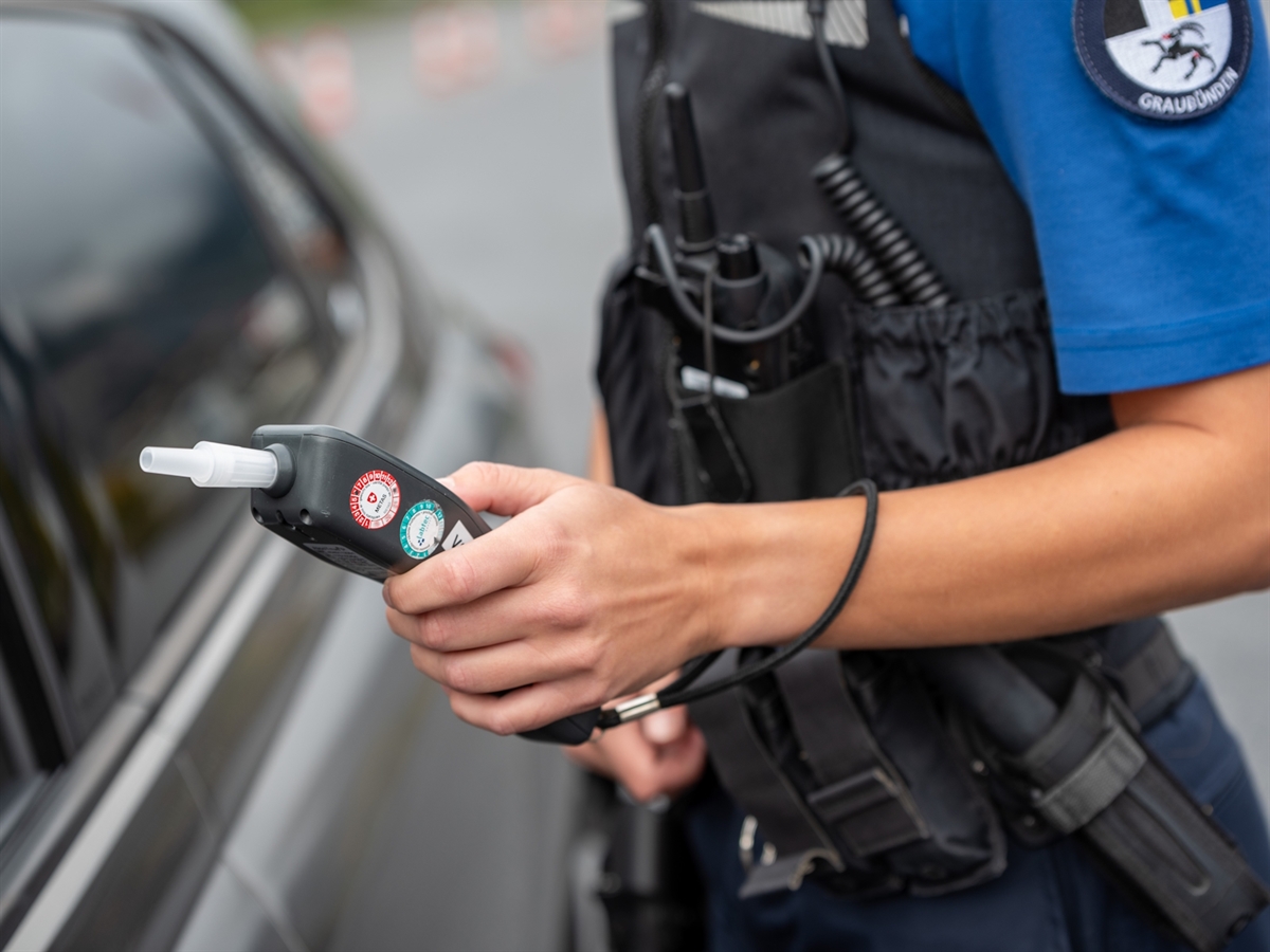 Eine Polizistin steht auf der Fahrerseite eines Autos und hält ein Atemalkoholmessgerät zum offenen Fenster der Fahrertüre hin.
