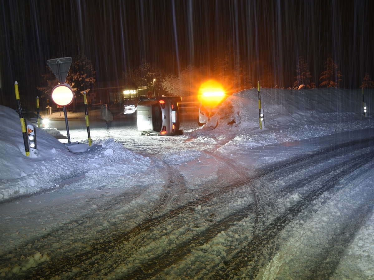 Die verschneite Landschaft der Ghiffakurve. Neben dem Erdwall liegt das Unfallauto auf der Seite. Zwei Polizeiautos, eines davon mit Blinklicht, erhellen die Nacht.