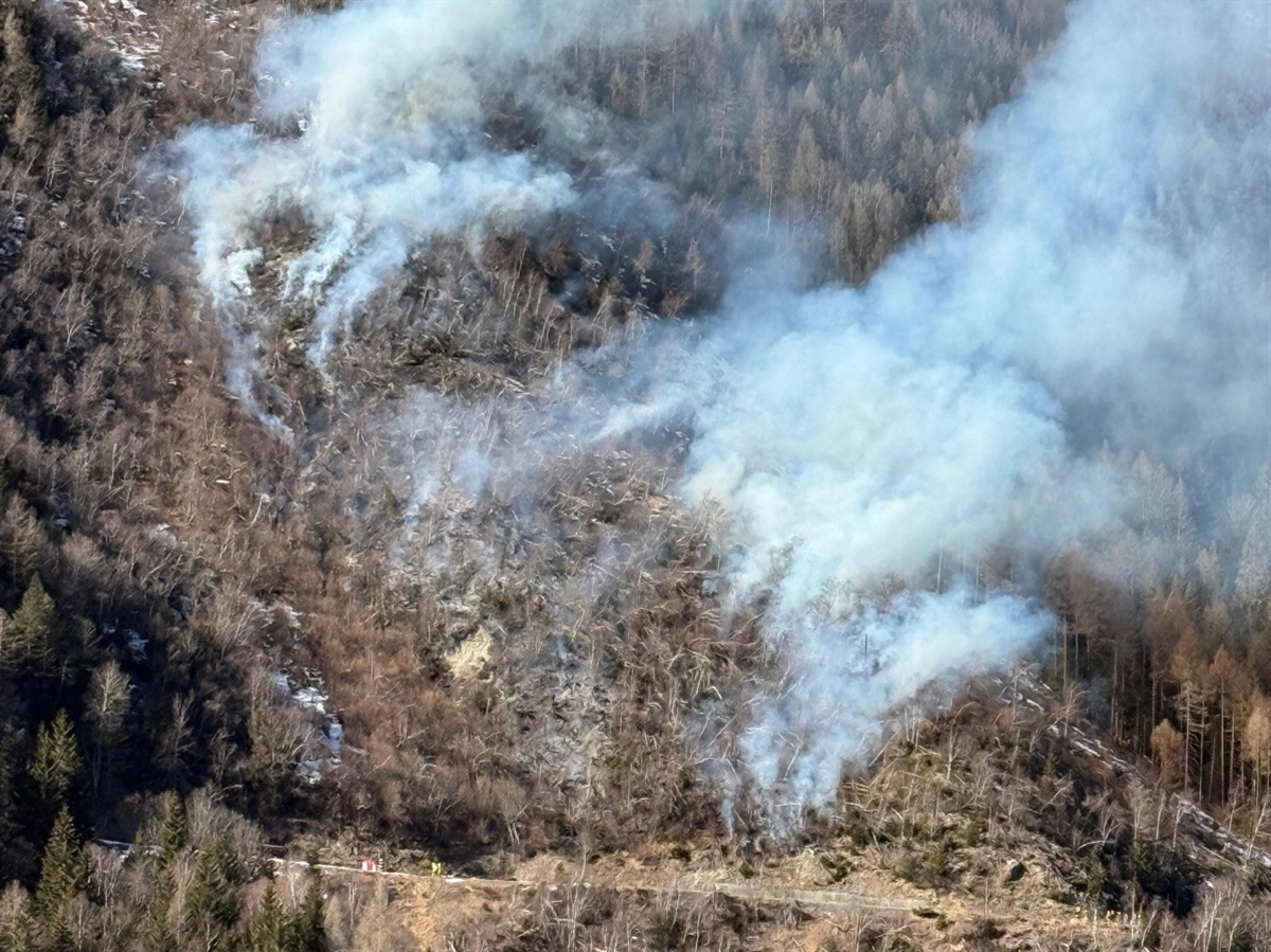 Blick vom Gegenhang auf das ansteigende Gelände, aus welchem Rauch emporsteigt.
